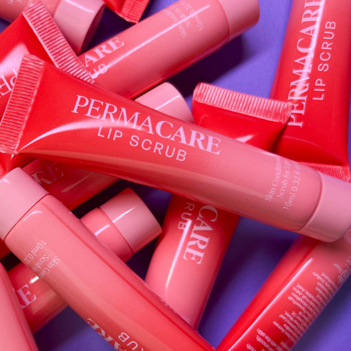 Plusieurs tubes de « Baume lèvres Permacare - 20 x 10 ml » dans les tons rose et rouge sont dispersés sur une surface violette, le nom du produit étant clairement visible sur chaque tube.